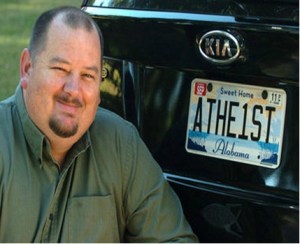 Photo of a man sitting next to his car, which has a license plate that reads, "ATHE1ST."