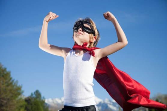 Picture of a boy wearing a Super Man cape, holding his arms up triumphantly and looking confidently at the sky